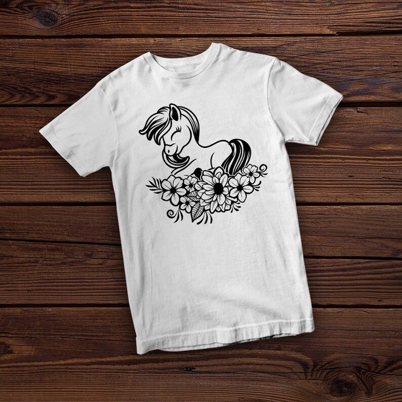 May include: White t-shirt featuring a black illustration of a horse lying on a bed of flowers. The horse has a flowing mane and tail, and the flowers include daisies and other floral elements. The design is centered on the shirt.