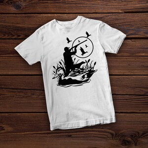 May include: White t-shirt featuring a black silhouette of a hunter aiming a rifle at flying ducks. The design includes a duck in the water and reeds, set against a circular backdrop. A hunting-themed graphic tee.