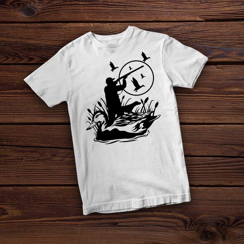 May include: White t-shirt featuring a black silhouette of a hunter aiming a rifle at flying ducks. The design includes a duck in the water and reeds, set against a circular backdrop. A hunting-themed graphic tee.