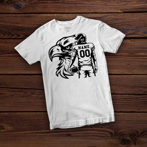 May include: White t-shirt with a black and white graphic of a football player holding a football and an eagle. The player's jersey has the text "NAME" and the number "00".