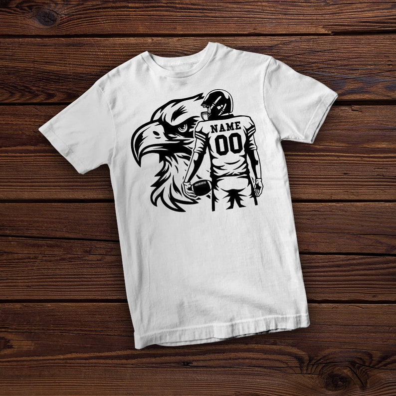 May include: White t-shirt with a black and white graphic of a football player holding a football and an eagle. The player's jersey has the text "NAME" and the number "00".