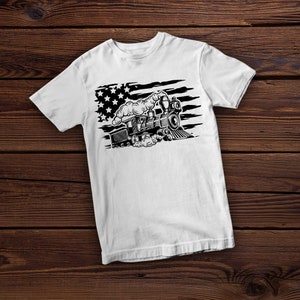 May include: White t-shirt featuring a black and white illustration of a vintage steam train. The design incorporates elements of the American flag, with stars and stripes. The train is depicted with detailed features, including smoke billowing from the smokestack.