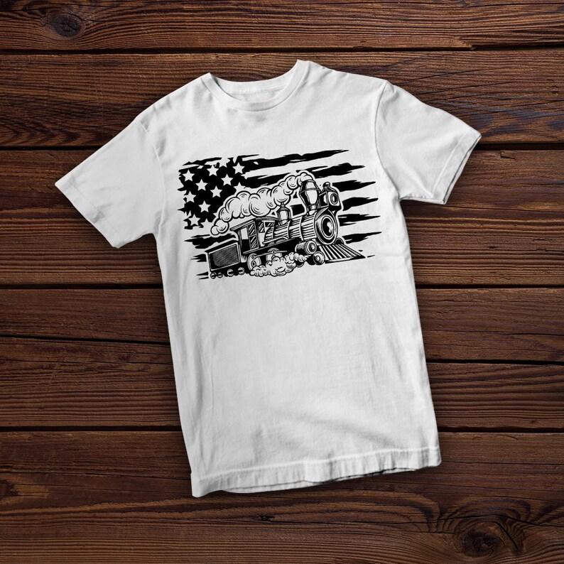 May include: White t-shirt featuring a black and white illustration of a vintage steam train. The design incorporates elements of the American flag, with stars and stripes. The train is depicted with detailed features, including smoke billowing from the smokestack.