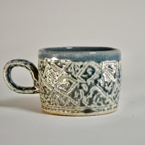 May include: A ceramic mug with a textured, geometric pattern in shades of blue, grey, and white. The mug has a rounded handle and a blue rim. The mug is approximately 7.6 cm tall.