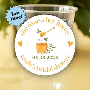 May include: A clear plastic cup featuring a round white sticker with the text "she found her honey" and "emily's bridal shower" in gold, a honey jar illustration with two bees, a honey dipper, green leaves, and the date "08.08.202X".
