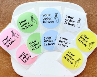 May include: A collection of circular stickers in various colors including light green, light blue, white, pink, and yellow. Each sticker features the black text your order is here and a line art illustration of a hand holding a small flower.