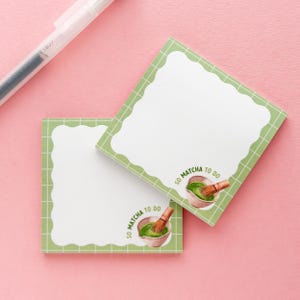 May include: Two square sticky notes with a white scalloped border and a green grid pattern. Each note features a watercolor illustration of matcha and the text "SO MATCHA TO DO". A white pen rests in the upper left corner.