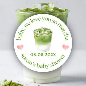 May include: A refreshing matcha drink in a clear plastic cup, topped with ice cubes. A white sticker on the cup reads "baby, we love you so matcha" with a watercolor illustration of a matcha drink, two pink hearts, and the text "susan's baby shower".