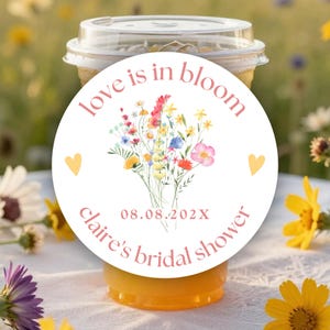 May include: A round white sticker on a clear plastic cup featuring a watercolor wildflower bouquet, two yellow hearts, and pink text that reads love is in bloom, 08.08.202X, and claire's bridal shower in a simple, clean, and legible font style.