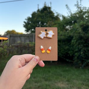 Può includere: Orecchini a tema Halloween su una scheda marrone. Gli orecchini presentano disegni di fantasmi e caramelle di mais. Gli orecchini a fantasma sono bianchi con un contorno giallo e gli orecchini a caramella di mais sono gialli, arancioni e bianchi.