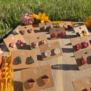 May include: A collection of small stud earrings displayed on brown cardboard cards, featuring autumn-themed shapes including pumpkins, acorns, mushrooms, maple leaves, coffee cups, and candy corn in various colors like orange, red, green, and white.