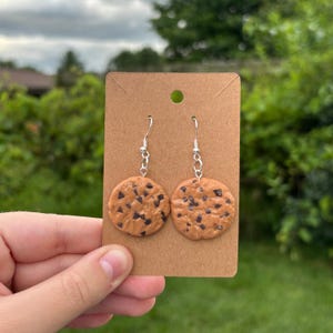 Puede incluir: Un par de pendientes novedosos con forma de galletas con chispas de chocolate. Los pendientes son redondos, con una base marrón claro y detalles de chispas de chocolate marrón oscuro. Cuelgan de ganchos plateados.