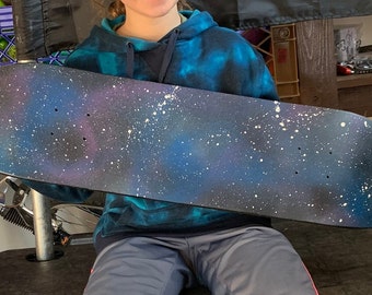 May include: A black skateboard deck painted with a galaxy design. The deck has a dark blue background with white and purple accents, and white speckles to represent stars.