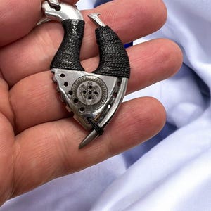 May include: A silver and black metal hook pendant with a detailed turtle design. The hook is wrapped in black cord and has a silver chain attached.