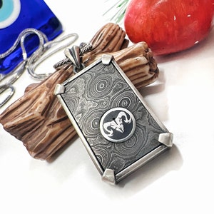 Könnte beinhalten: Eine silberne Halskette mit einem rechteckigen Anhänger, der ein schwarzes und weißes Widderkopf-Design zeigt. Der Anhänger hat eine strukturierte Oberfläche mit einem kreisförmigen Muster.