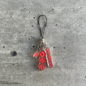 May include: A clear acrylic charm with a black strap. The charm features three red circles and a rectangular tag with the word "ARIRANG" in red. The charm is attached to a silver metal chain.