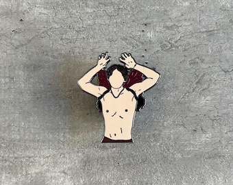 May include: A silver enamel pin depicting a person with their arms raised above their head, wearing a red shirt. The person is bare-chested and has short, dark hair.