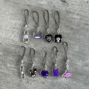 Może przedstawiać: Dziewięć różnych charmów do telefonu z czarnym sznurkiem i przezroczystymi akrylowymi charmów. Charmy prezentują różne wzory, w tym rybę z napisem "SUPER", serce z kwiatowym wzorem, osobę w czarnym kapeluszu i czerwonym nosem, koło z wieloma mniejszymi kółkami, serce z czarno-białą twarzą, serce z literami "JK", fioletową sylwetkę litery "B" i wieloryba z różowym ciałem.