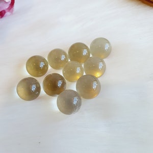 May include: A collection of translucent, spherical gemstones in varying shades of yellow and brown. The polished spheres are scattered on a white, fluffy surface, with light reflecting off their smooth surfaces. The gemstones appear to be approximately 2.5 cm in diameter.
