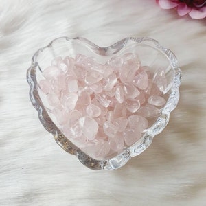 May include: A clear, heart-shaped glass bowl filled with small, polished rose quartz stones. The stones are a soft pink colour. The bowl sits on a white, fluffy surface, with pink flowers in the background.