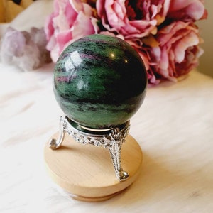 May include: A green and pink gemstone sphere, displayed on a silver-coloured metal stand with decorative features. The stand is placed on a round, light-coloured wooden base. A bouquet of pink flowers is in the background.