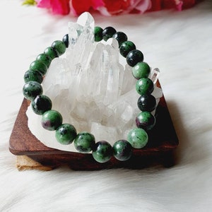 May include: A gemstone bracelet with green and red speckled beads. The bracelet is displayed on a small wooden stand, set against a backdrop of clear quartz crystals. Pink flowers are visible in the background.