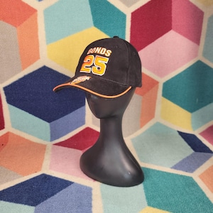 Könnte beinhalten: Schwarze Baseballkappe mit dem Wort "BONDS" und der Zahl "25" in orangefarbenen und weißen Buchstaben. Die Kappe hat einen orangefarbenen Schirm und wird auf einem schwarzen Schaufensterkopf präsentiert.