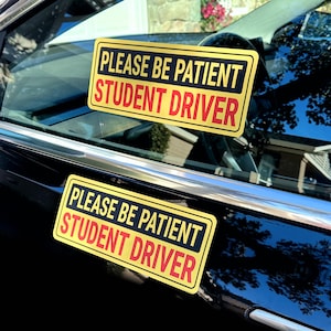 May include: Two yellow rectangular signs with black borders and text that reads "PLEASE BE PATIENT STUDENT DRIVER" in black and red. The signs are affixed to the window of a black car.