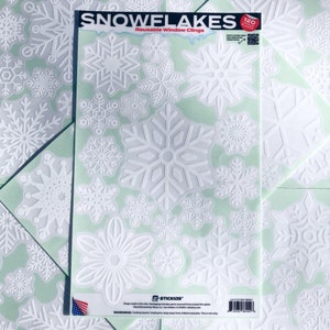 May include: A package of white snowflake window clings. The package reads "SNOWFLAKES" and "Reusable Window Clings". The clings are various sizes and designs. The background is a light green color with additional snowflake clings.