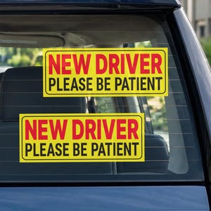 May include: Two rectangular yellow car decals applied to a rear vehicle window. Each sticker features the text NEW DRIVER in bold red capital letters, followed by the text PLEASE BE PATIENT in bold black capital letters on a yellow background.