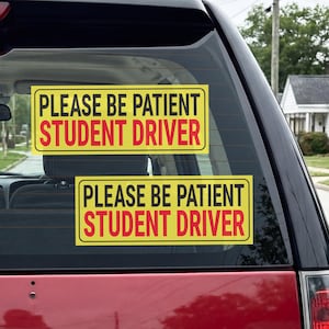 May include: Two rectangular yellow car decals applied to the rear window of a red vehicle. Each decal features a black border and the text PLEASE BE PATIENT in black, followed by the words STUDENT DRIVER in bold red capital letters.