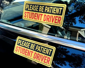 May include: Two rectangular yellow car decals with a black border. The text reads PLEASE BE PATIENT in black capital letters on the top line and STUDENT DRIVER in red capital letters on the bottom line. One decal is on the window and one is on the door.