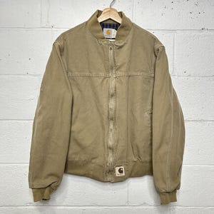 May include: A tan canvas bomber jacket with a full-length front zipper, ribbed collar, and ribbed cuffs. The jacket features a dark blue and green plaid interior lining, a Carhartt brand label on the collar, and a square Carhartt patch at the hem.