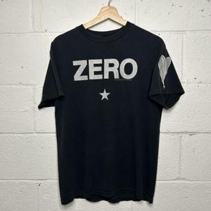 May include: A black short-sleeve t-shirt featuring the word ZERO printed in large white capital letters across the chest. Below the text is a small white star graphic. The right sleeve displays a white heart-shaped design with a trailing line.