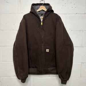 Puede incluir: Una chaqueta con capucha de color marrón oscuro con cremallera frontal completa, mangas largas y puños acanalados. La chaqueta presenta un forro interior gris, una capucha con cordón y una pequeña etiqueta rectangular de la marca Carhartt.