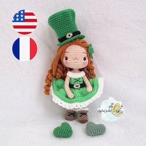Peut inclure: Une poupée en crochet portant une robe verte, un chapeau vert avec un bandeau noir et des bottes brunes. La poupée a de longs cheveux bruns bouclés et un trèfle vert sur sa robe. La poupée est assise sur une surface blanche avec deux cœurs verts.