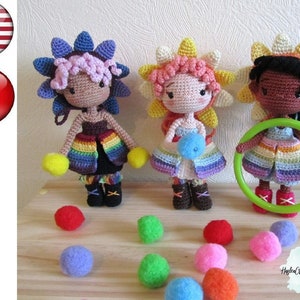 May include: Three crocheted dolls with rainbow skirts and flower headbands. The dolls are standing on a wooden surface with colorful balls scattered around them.