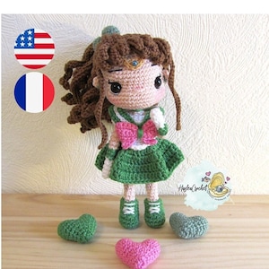 Puede incluir: Muñeca de crochet con cabello castaño, vestido verde y lazo rosa. La muñeca lleva zapatos blancos y está de pie sobre una superficie de madera con corazones verdes y rosas.