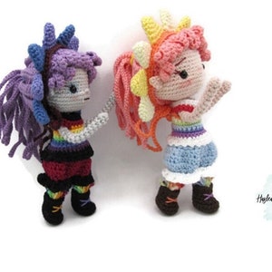 May include: Two crocheted dolls with rainbow striped tops and colorful hair. The doll on the left has purple hair and black pants. The doll on the right has pink and yellow hair and blue pants.