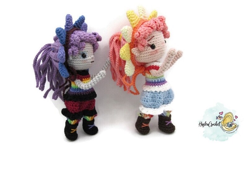 May include: Two crocheted dolls with rainbow striped tops and colorful hair. The doll on the left has purple hair and black pants. The doll on the right has pink and yellow hair and blue pants.