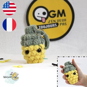 May include: A crocheted pineapple grenade keychain with a green top and yellow body. The keychain has a small key ring attached. The keychain is sitting on a white surface with a yellow and black circle sticker that says "OGM J'EN VEUX TOUJOURS PAS".