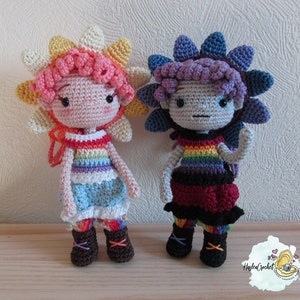 May include: Two crocheted dolls with rainbow-colored clothing and flower-shaped hats. The doll on the left has a pink and yellow hat and a blue dress. The doll on the right has a purple and blue hat and a red dress.
