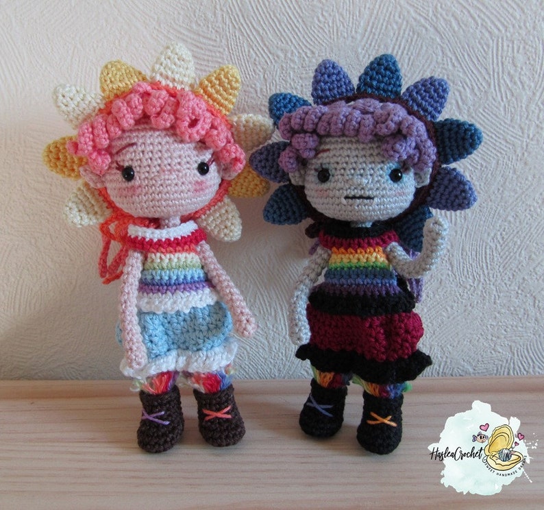 May include: Two crocheted dolls with rainbow-colored clothing and flower-shaped hats. The doll on the left has a pink and yellow hat and a blue dress. The doll on the right has a purple and blue hat and a red dress.