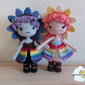 May include: Two crocheted dolls with rainbow skirts and flower-shaped hats. One doll has a purple hat and a blue and purple skirt, while the other doll has a yellow hat and a blue and orange skirt.