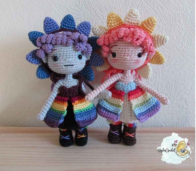 May include: Two crocheted dolls with rainbow skirts and flower-shaped hats. One doll has a purple hat and a blue and purple skirt, while the other doll has a yellow hat and a blue and orange skirt.