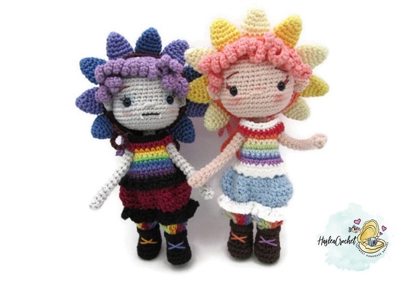 May include: Two crocheted dolls with rainbow striped tops and flower-shaped hair. The doll on the left has purple hair and a red skirt. The doll on the right has pink hair and a blue skirt.
