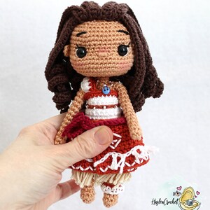 May include: Crocheted doll with brown hair, wearing a red and white dress with a brown and white skirt. The doll has a blue button on its chest.