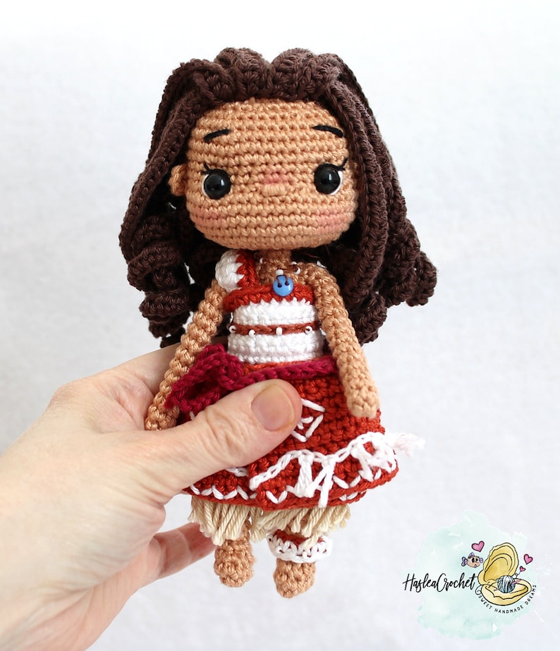 May include: Crocheted doll with brown hair, wearing a red and white dress with a brown and white skirt. The doll has a blue button on its chest.