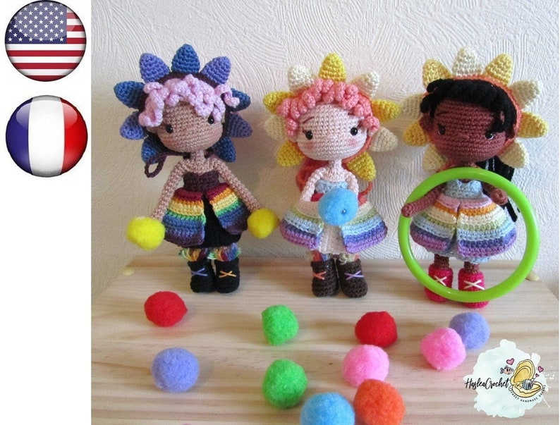 May include: Three crocheted dolls with rainbow skirts and flower headbands. The dolls are standing on a wooden surface with colorful balls scattered around them.