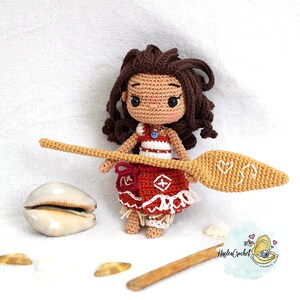 May include: A crocheted doll of a young girl with brown hair, wearing a red and white dress and holding a wooden paddle. The doll is standing on a white surface with seashells.